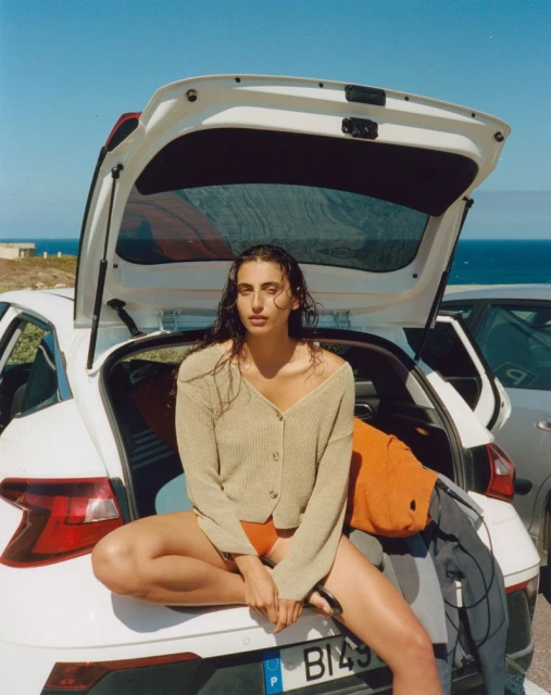 Woman poses seated on a white car's trunk with the open hatch, ocean and blue sky background, wearing a sweater and swimsuit.