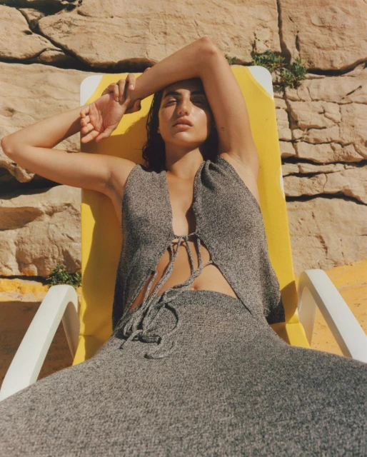 A woman in grey knitwear reclines on a yellow deck chair with a stone wall background on a sunny day, creating a relaxed and fashionable outdoor scene.