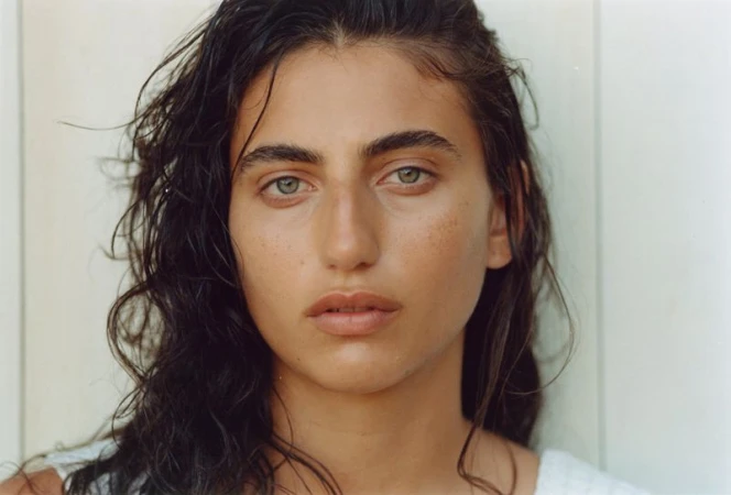 Close-up portrait of a young adult female with light skin, green eyes, and dark, wet wavy hair against a white wall background. 
