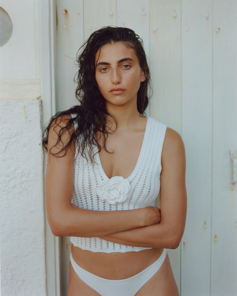 Portrait of a young adult female wearing a white crocheted top and white bottoms, with arms crossed in front of a white wall.