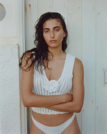 Portrait of a young adult female wearing a white crocheted top and white bottoms, with arms crossed in front of a white wall.