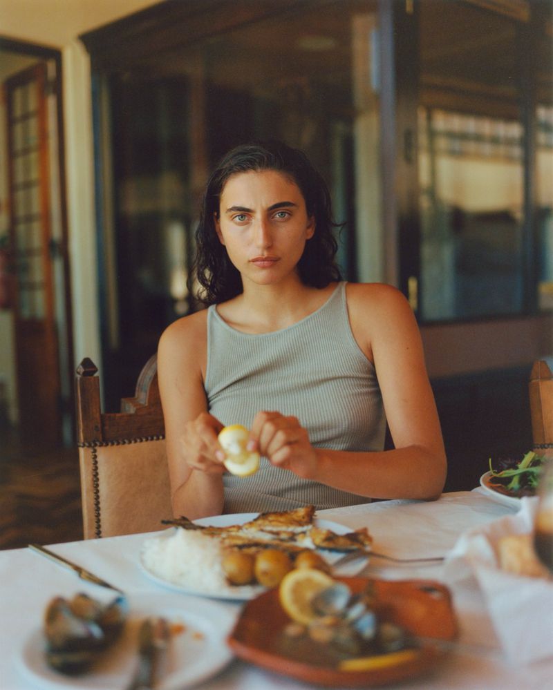 Young woman with dark hair is sitting at a table in an indoor setting preparing a meal of fish, rice, and vegetables.