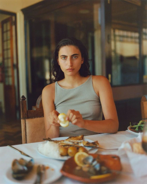 Young woman with dark hair is sitting at a table in an indoor setting preparing a meal of fish, rice, and vegetables.