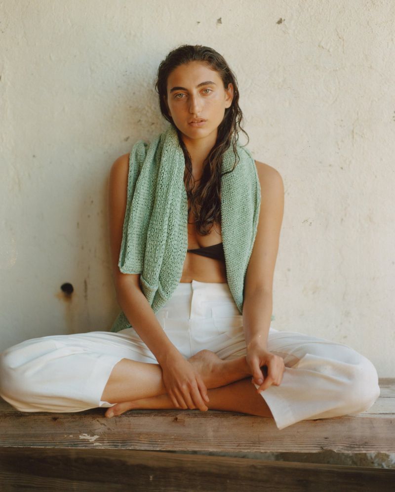 Woman in white pants and a light green top sits cross-legged on a wooden surface in front of a textured wall, natural light.