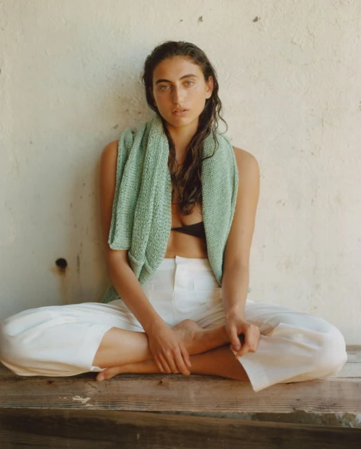 Woman in white pants and a light green top sits cross-legged on a wooden surface in front of a textured wall, natural light.