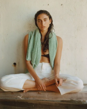 Woman in white pants and a light green top sits cross-legged on a wooden surface in front of a textured wall, natural light.