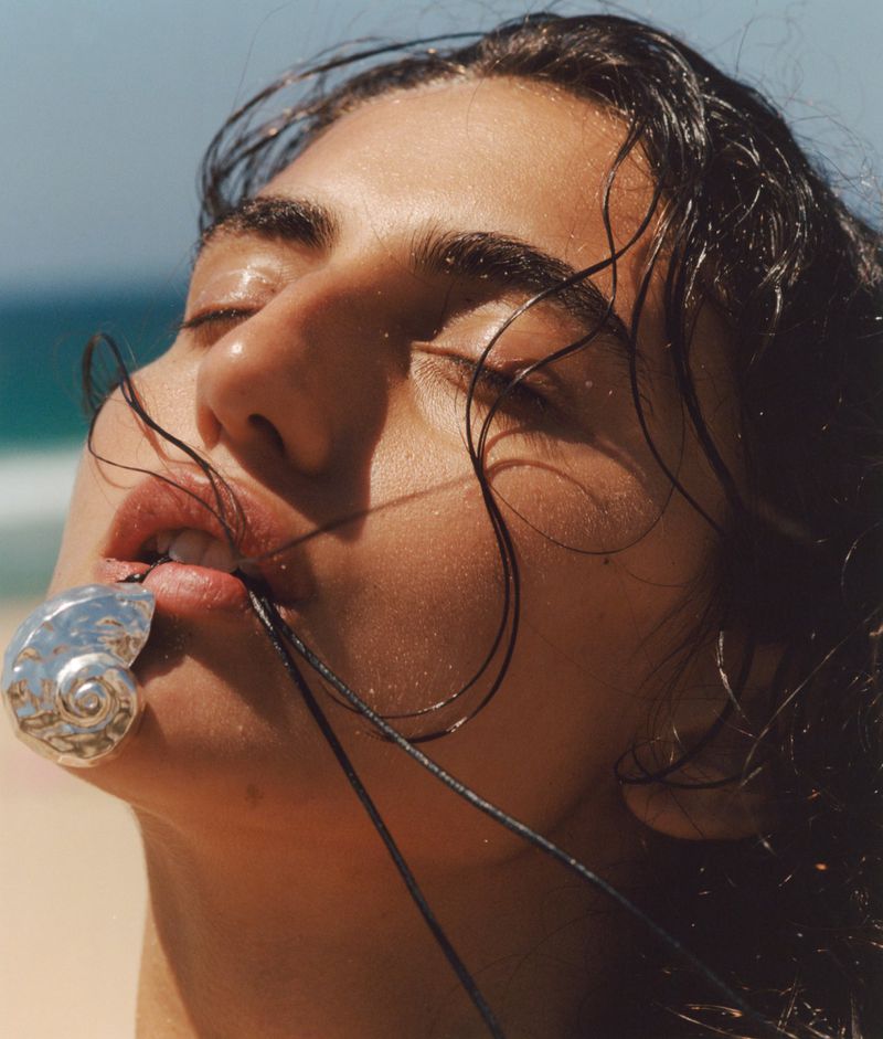 Close-up of a woman with wet hair wearing a shell necklace on a beach under a blue sky, with light tan sand and ocean view.