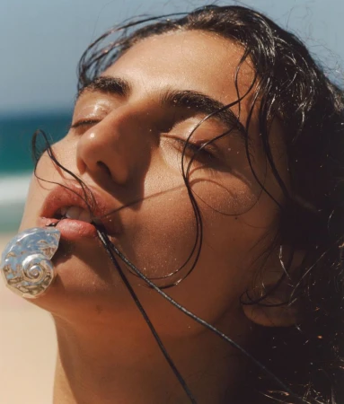 Close-up of a woman with wet hair wearing a shell necklace on a beach under a blue sky, with light tan sand and ocean view.