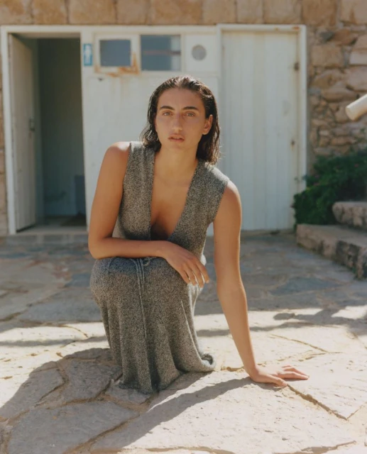 Adult woman with wet hair poses in a sleeveless dress, crouching in front of a white building with stone exterior on a sunny day.
