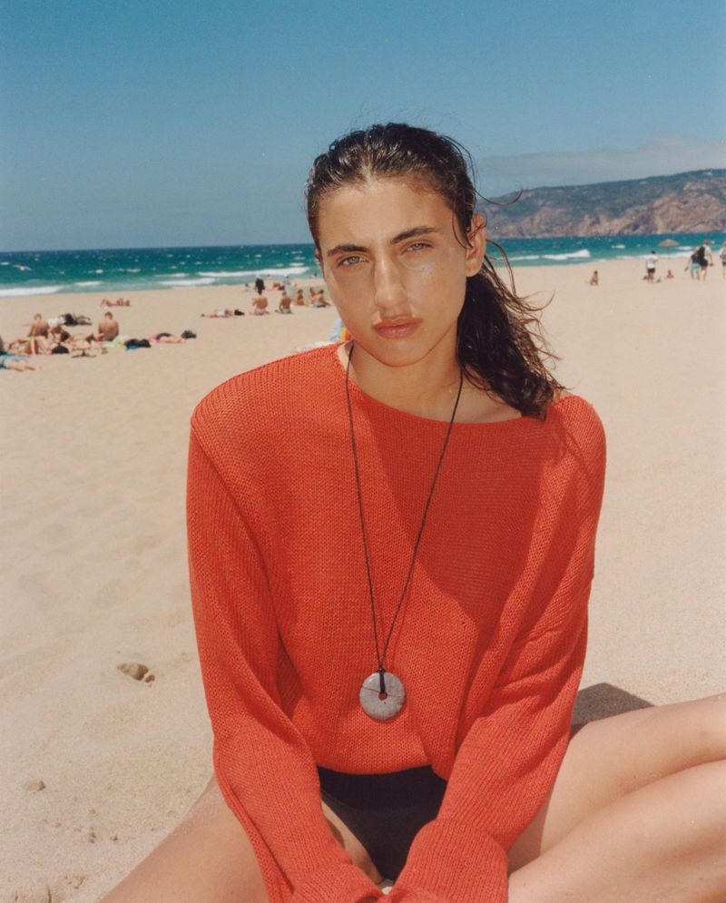 Young woman with red top poses on beach, bathed in sunlight with ocean background, showcasing fashion and relaxed, outdoor summer vibe.