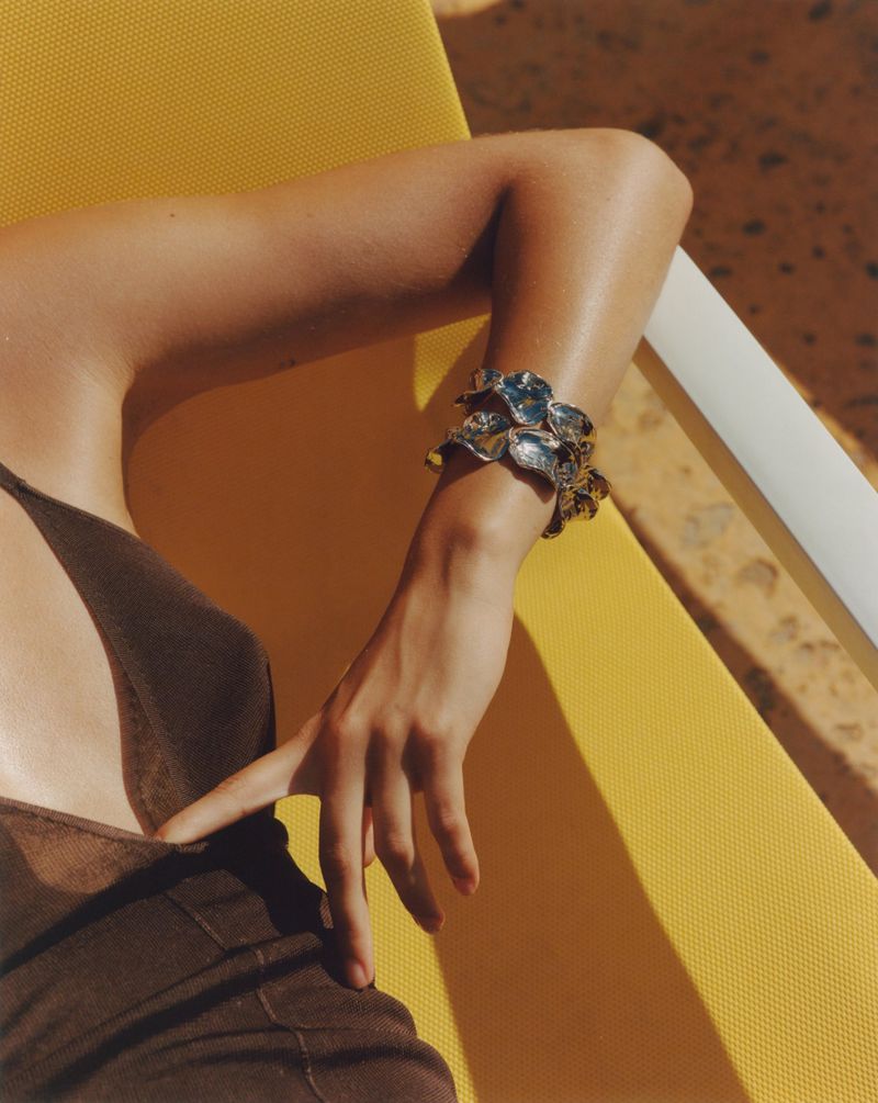 Close up of a woman's arm adorned with a silver cuff bracelet, resting on a yellow surface under bright, warm sunlight, creating strong shadows.