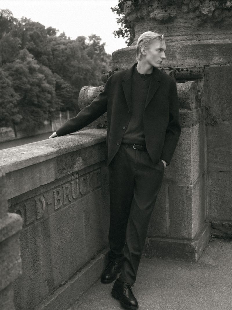 Blond male model in black suit poses by stone bridge with green trees in background in black and white for fashion editorial.