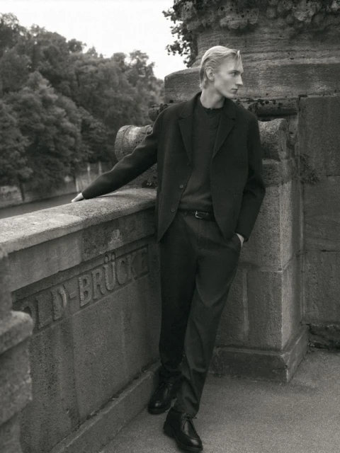 Blond male model in black suit poses by stone bridge with green trees in background in black and white for fashion editorial.