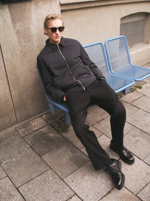 Blond male fashion model in sunglasses sits on a blue metal bench in an urban setting wearing a black jacket, pants, and dress shoes.