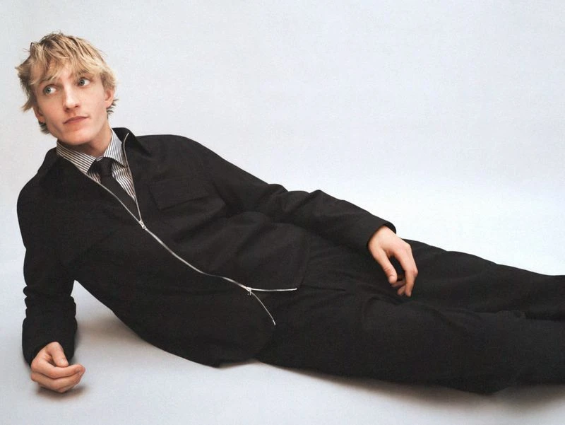 A young adult male with blond hair poses in a dark suit and tie while reclining on a light gray surface in a studio setting.
