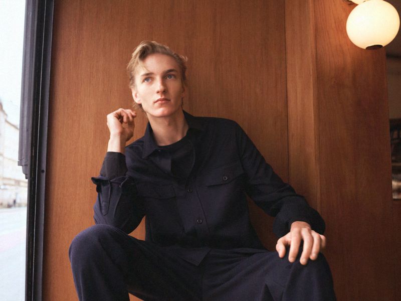 Blond male model poses indoors wearing a dark-blue button-down shirt and trousers, with a wood-panel wall and modern lighting in the background.
