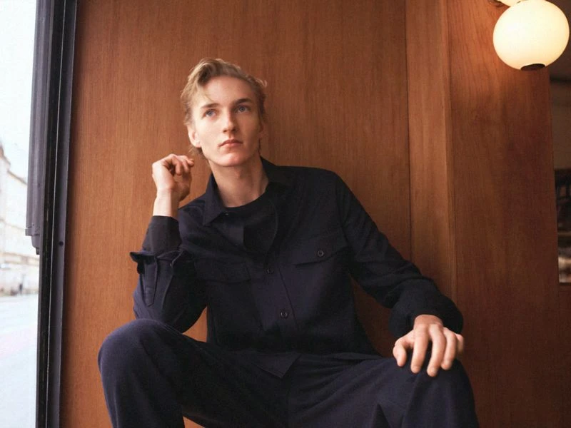 Blond male model poses indoors wearing a dark-blue button-down shirt and trousers, with a wood-panel wall and modern lighting in the background.