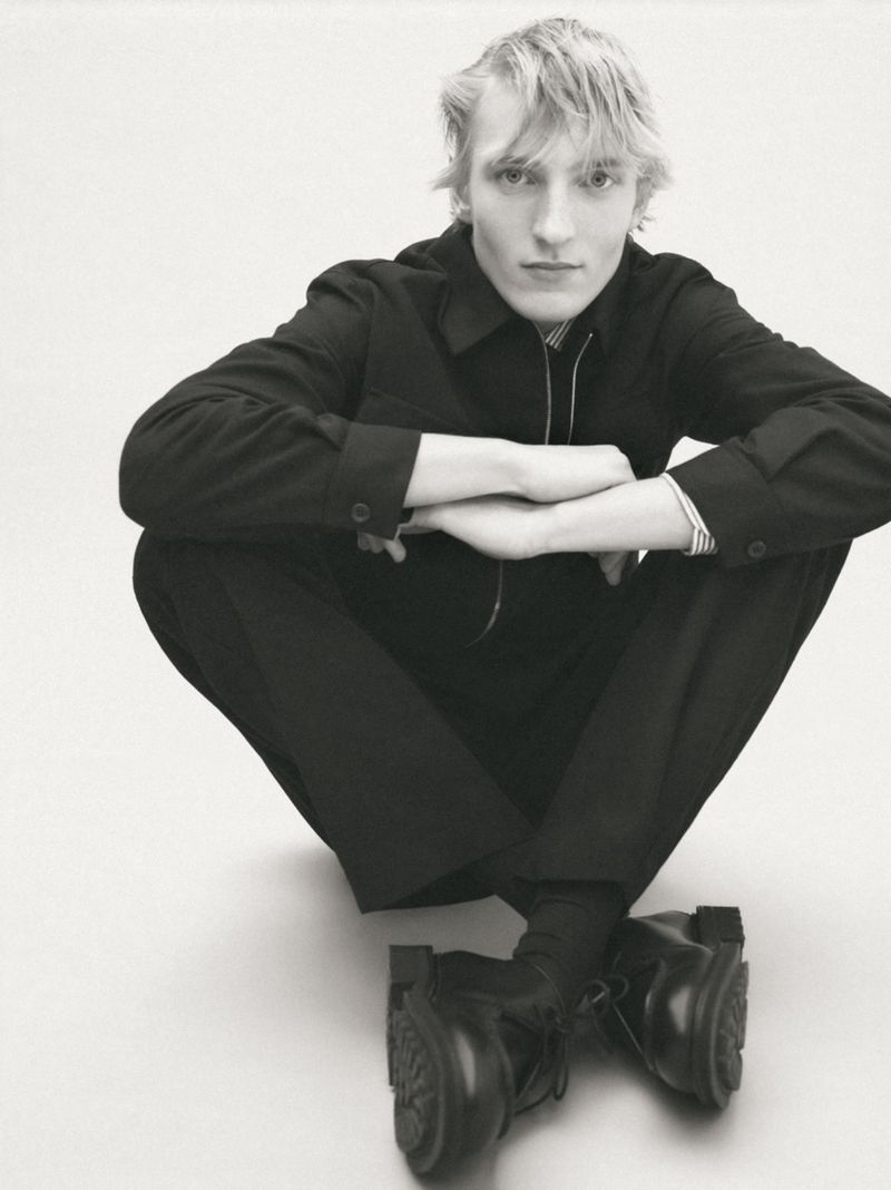 Young adult male model with blond hair poses in studio. He wears a black jacket, trousers, socks, and chunky lug-sole dress shoes, arms crossed.