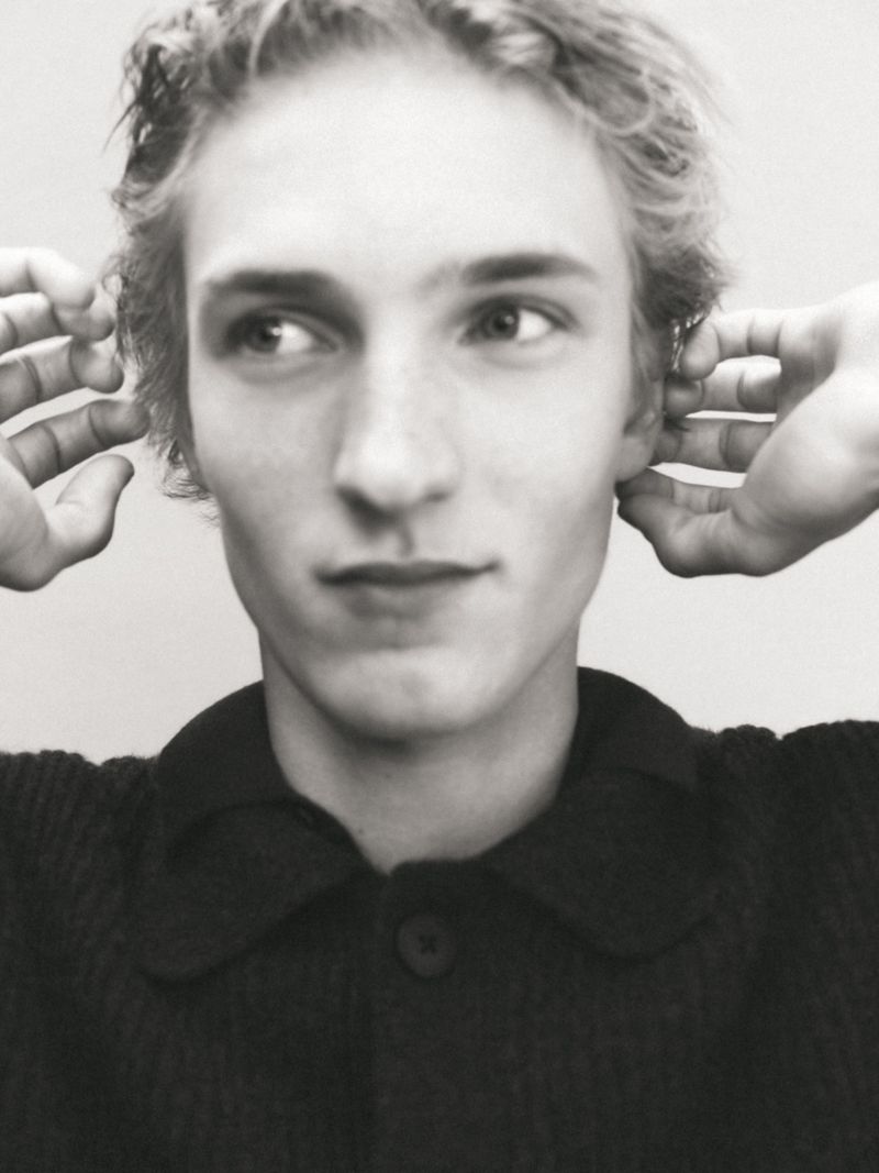 Close-up monochrome portrait of a young male model with hands framing his head against a plain background