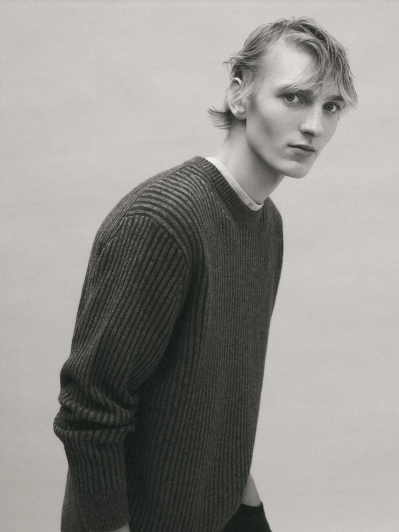 Monochrome portrait of a young adult male with light hair, wearing a dark knit sweater, posing against a plain grey background.