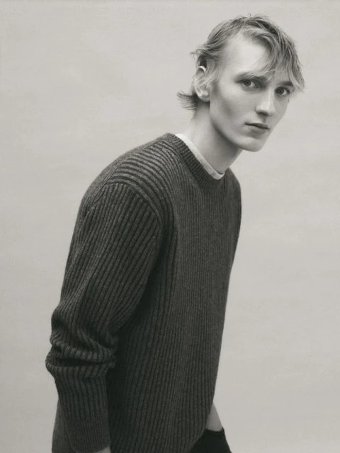 Monochrome portrait of a young adult male with light hair, wearing a dark knit sweater, posing against a plain grey background.