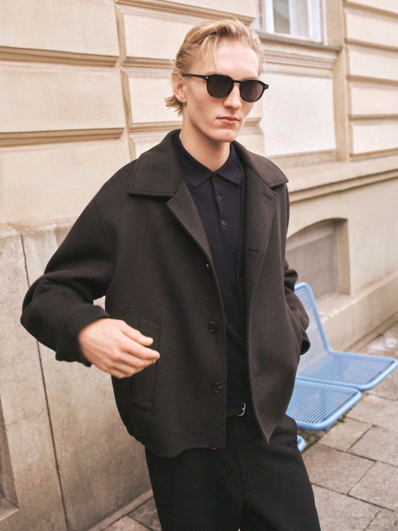 Man wearing sunglasses, a black jacket, and black trousers stands outside in an urban setting, posing for fashion or editorial photography.