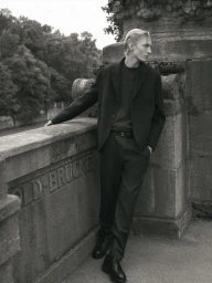 Monochrome fashion portrait of a male model leaning against a stone bridge wall in a dark jacket and trousers.