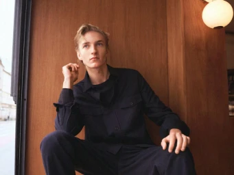 A young male model wearing a dark shirt and trousers seated indoors against a warm wooden panel wall.