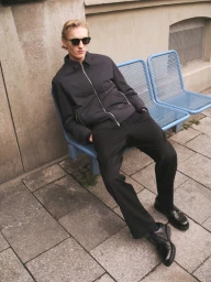 Man wearing sunglasses and a black jacket seated on a blue metal bench outdoors against a stone wall.