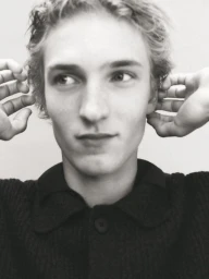 Close-up monochrome portrait of a young male model with hands framing his head against a plain background