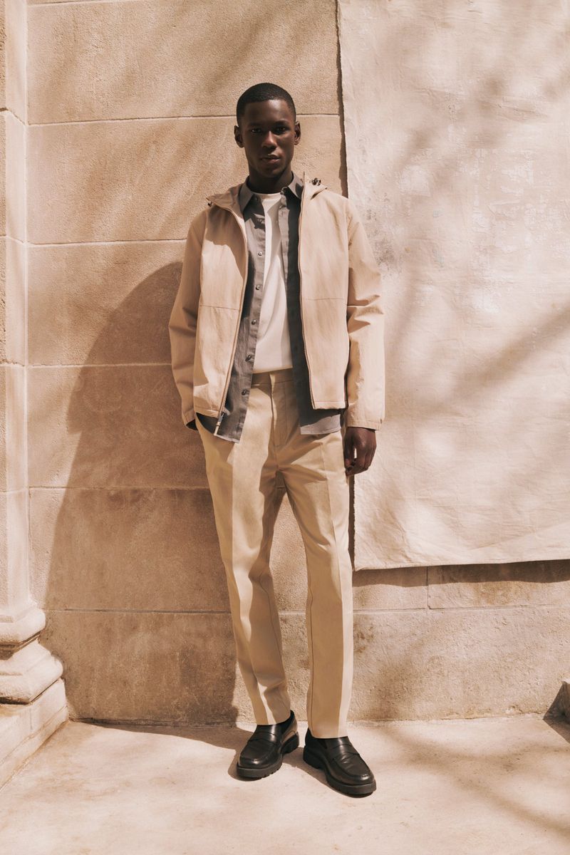 A full shot of a stylish adult male wearing a light hooded jacket, light shirt, tan trousers, and black shoes, posing against a wall.