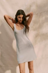 Model in a light grey slip dress posing with hands behind her head against a textured canvas backdrop