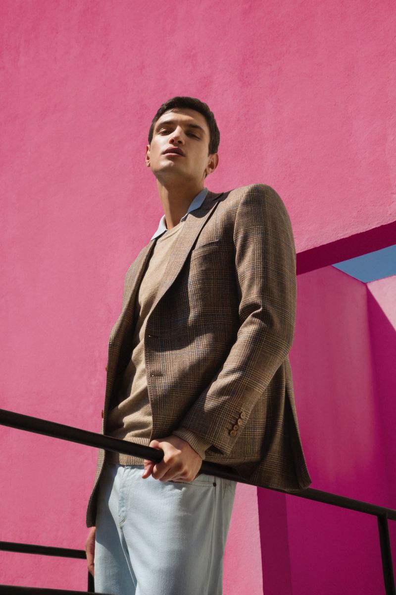 An adult male model poses in a blazer, sweater, and light blue pants against a pink wall and black railing, shot at a low angle.