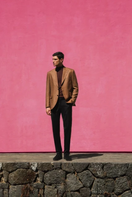 Man poses against pink wall wearing brown blazer and dark turtleneck, standing on a stone wall in natural, eye-level, medium shot.