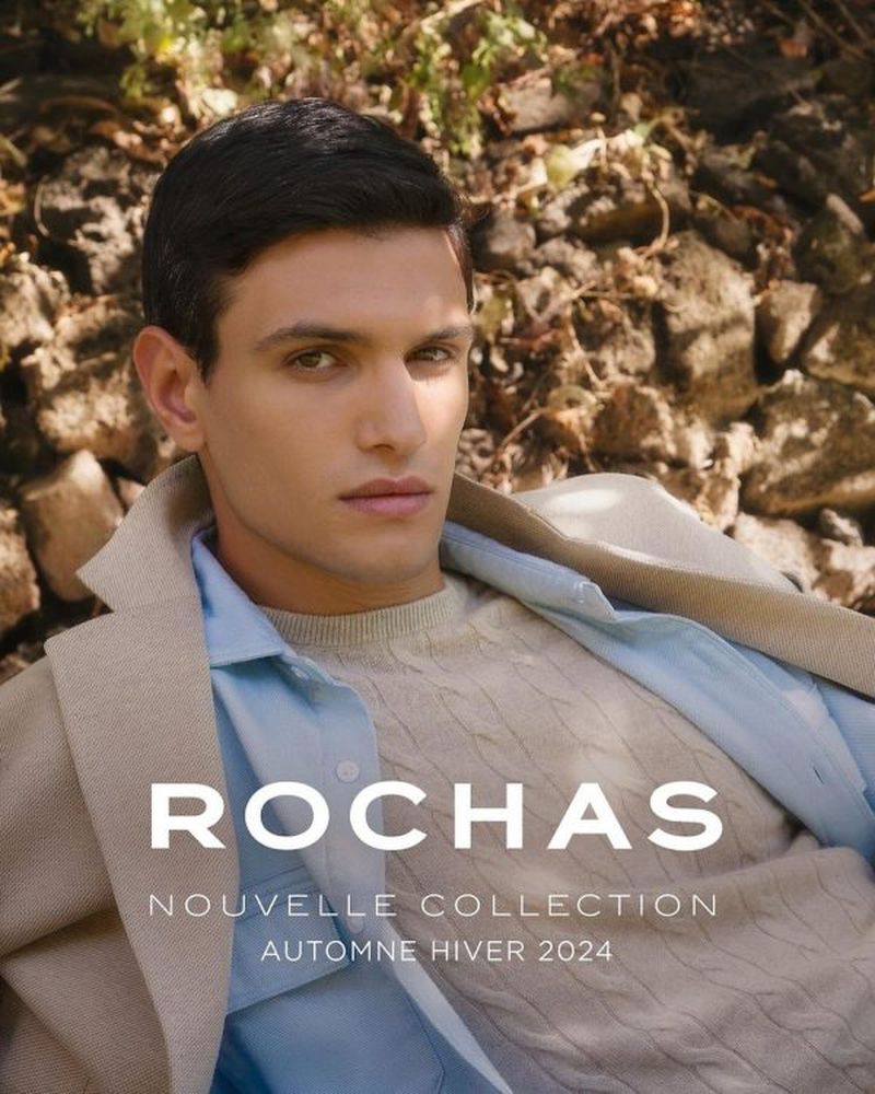 Man wearing a sweater, shirt, and coat poses outdoors against a rocky background for a ROCHAS Autumn/Winter 2024 collection ad.