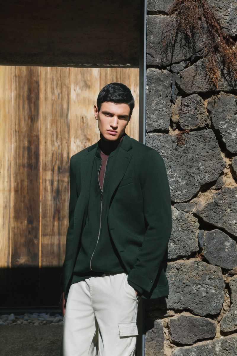 A stylish man wearing a green blazer stands next to a textured stone wall and wooden door in an outdoor setting with natural light.