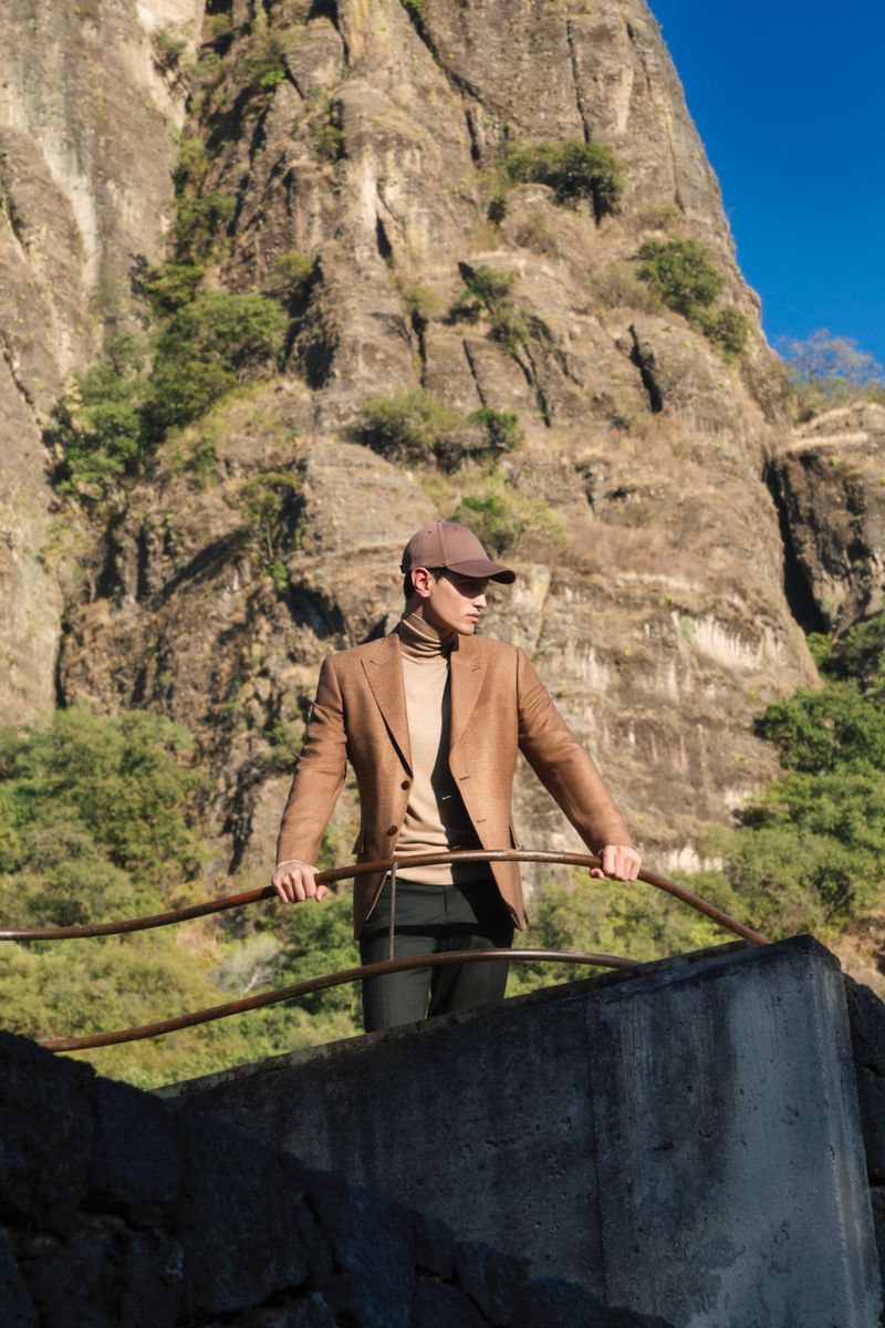 Man in a blazer, turtleneck, and cap poses on a concrete structure, with a mountain and blue sky as a backdrop on a sunny day.