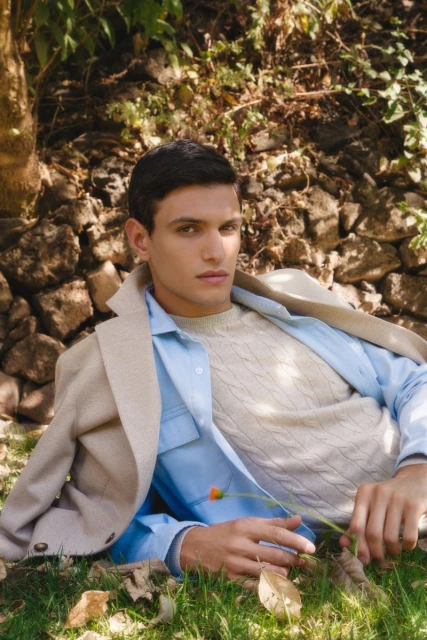 Adult male lying on grassy ground outside wearing a sweater, jacket, and dress shirt. He holds a small orange flower.