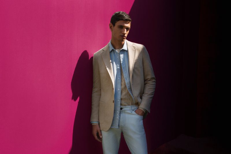 A handsome man poses in a beige suit jacket and light jeans against a solid magenta wall in a fashion or portrait photography style.