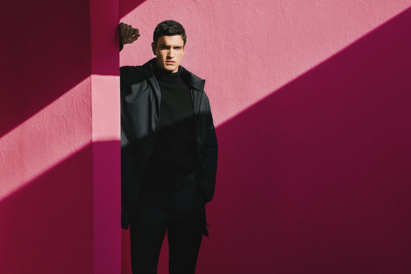 Stylish man in black turtleneck and coat poses against pink wall with angled sunlight, creating geometric shadow patterns and stark contrast.