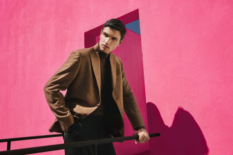 A young adult male poses in front of a vibrant pink wall, wearing a brown blazer and dark trousers, for a fashion-style outdoor photoshoot.
