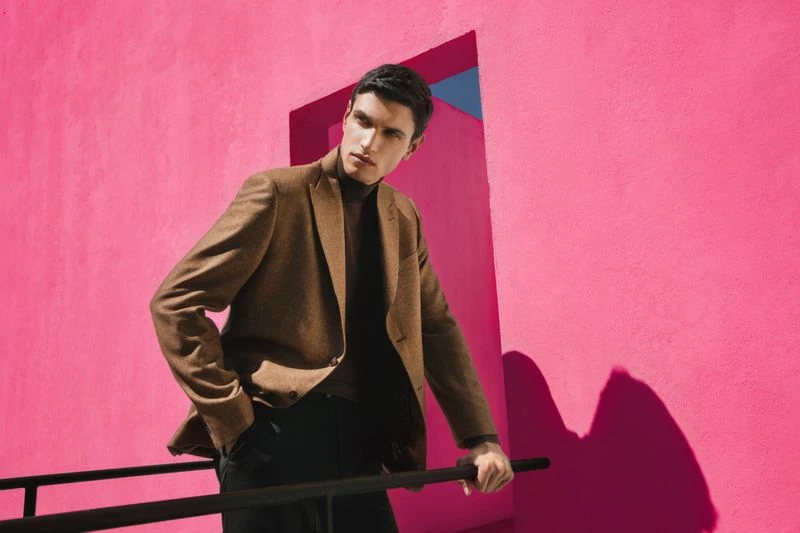 A young adult male poses in front of a vibrant pink wall, wearing a brown blazer and dark trousers, for a fashion-style outdoor photoshoot.