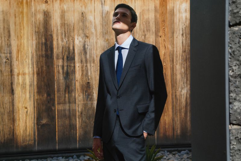 An adult man in a suit stands outdoors in dappled sunlight, looking up with a slight smile, wearing a suit, shirt, and tie.