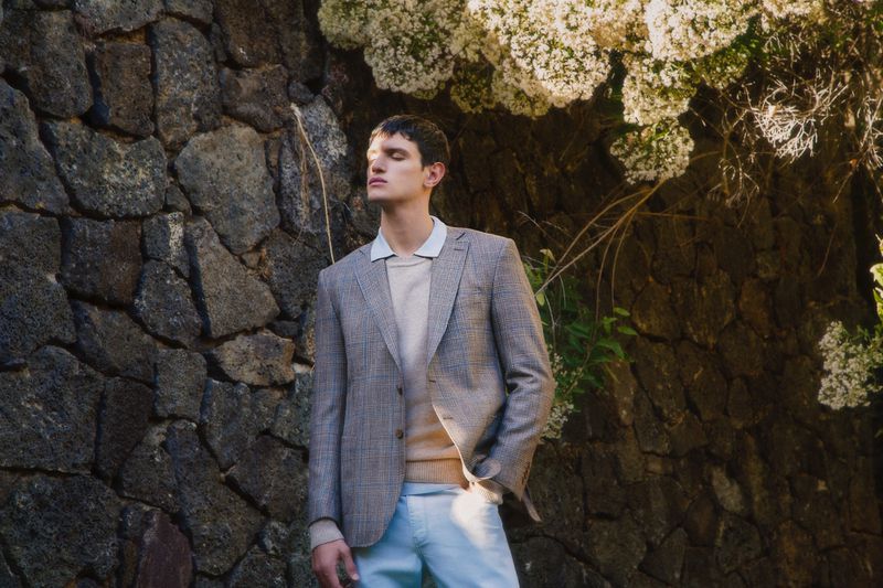 A young man in a blazer and light-colored trousers stands outdoors with his eyes closed, posing against a stone wall with flowering plants.
