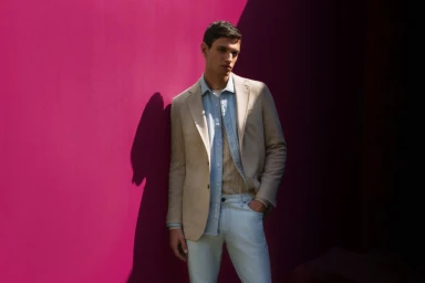 Male model wearing a beige blazer and light blue shirt standing against a vibrant pink wall.