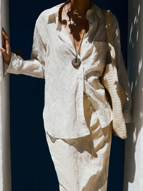 Woman in a beige collared shirt and skirt posing outdoors in front of a blue wall, wearing a long pendant necklace and a knit bag.