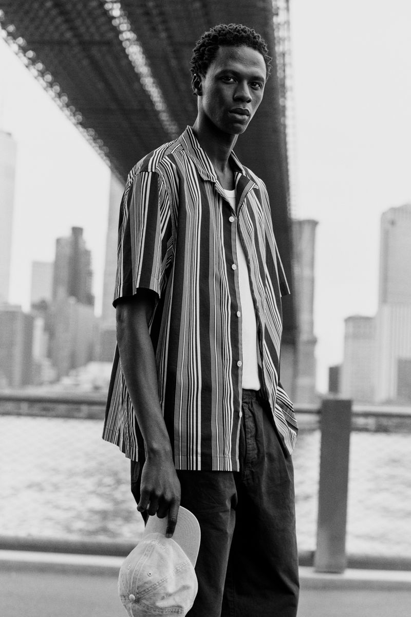 Black and white shot of a man in a striped shirt standing near the Brooklyn Bridge, holding a cap, with city buildings in the background.