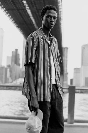 Black and white shot of a man in a striped shirt standing near the Brooklyn Bridge, holding a cap, with city buildings in the background.