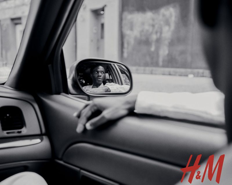 Black and white shot inside a car reflecting an adult male in the rearview mirror, showcasing a city building outside, H&M logo visible.