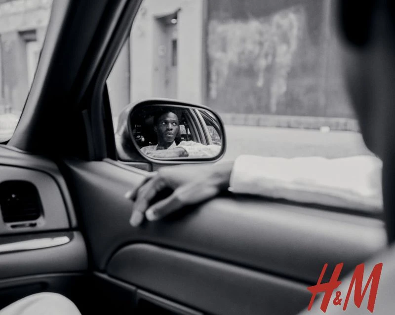 Black and white shot inside a car reflecting an adult male in the rearview mirror, showcasing a city building outside, H&M logo visible.